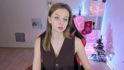 Media: Video of a young, fair-skinned woman with long, straight blonde hair, wearing a black sleeveless vest, sitting in a gaming chair in a modern, brightly lit room with neon decorations.