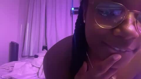 Media: A video of a Black woman with medium-brown skin, wearing glasses, a black hair tie, and headphones, touching her neck in a dimly lit room with purple lighting and white curtains.