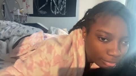 Media: Video of a young Black woman with braided hair, wearing a tie-dye shirt, lying on a bed with a white and gray comforter. Background features a wall with a black and white abstract painting and a white metal shelf.