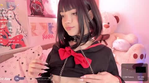 Video of a young woman with a bob haircut, wearing a black dress with a red bow and chain collar, in a cozy, pink-themed room with plush toys and anime posters.