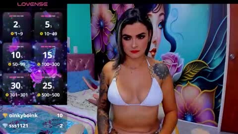 Media: Video of a tattooed Latina woman in a white bikini, standing in a colorful room with a vibrant floral mural.