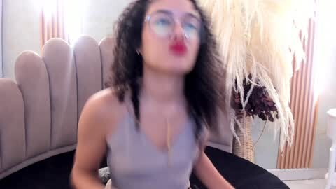 Media: Video of a woman with curly hair, wearing a low-cut gray top, sitting on a curved beige couch, making a kissy face; background features beige walls with decorative elements.