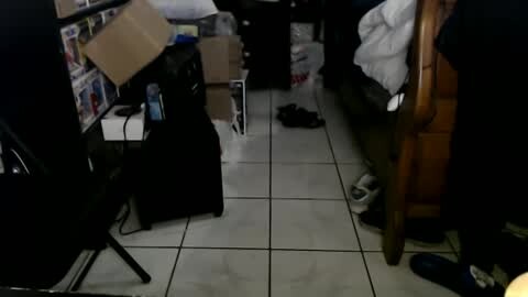 Media: Video of a cluttered, dimly lit bedroom with a wooden bed, black suitcases, and scattered clothing on a white tiled floor.