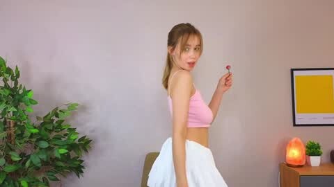 Media: Video of a slim, light-skinned woman with blonde hair, wearing a pink sports bra and white skirt, holding a red lollipop, in a minimalist room with a green plant, wooden desk, and a framed picture.