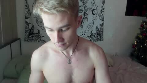 Media: Video of a young, shirtless, fair-skinned man with short, blonde hair, wearing a necklace, standing in a bedroom with a black and white tapestry on the wall and a decorated Christmas tree in the background.