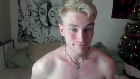 Media: Video of a shirtless, light-skinned, young man with short, blonde hair, wearing a necklace, in a bedroom with a tapestry, Christmas tree, and bed.
