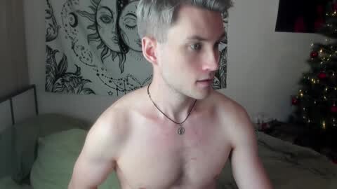 Media: Video of a shirtless, light-skinned, young man with short, dyed blonde hair, wearing a silver necklace, in a dimly lit bedroom with a green bedspread, black tapestry, and a Christmas tree.