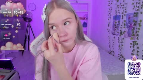Media: Video of a young woman with long, straight blonde hair and light skin, wearing a pink t-shirt, applying makeup while sitting in a dimly lit bedroom with purple lighting.