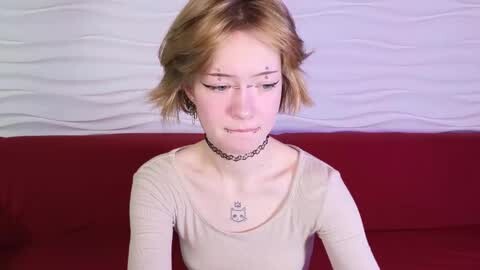 Media: Video of a young woman with short, light brown hair, wearing a beige top, black choker, and makeup. She sits on a red couch against a textured white wall.