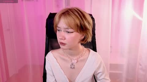 Media: Video of a young, fair-skinned woman with short, light brown hair, wearing a white, off-shoulder top, black necklace, sitting in a black chair. Pink and white sheer curtains in the background create a soft, romantic ambiance.