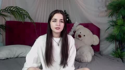 Media: Video of a young woman with long dark hair, wearing a white sweater, sitting on a bed with a red headboard and a large teddy bear.