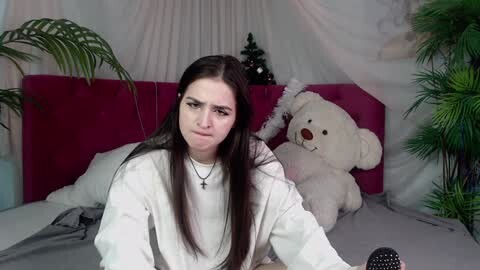 Media: A video of a young woman with long dark hair, wearing a white sweater, sitting on a bed with a plush white teddy bear and a Christmas tree in the background.