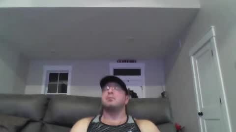 Media: Video of a man with glasses and a black cap, wearing a grey tank top, sitting on a grey couch in a modern, white-walled living room.