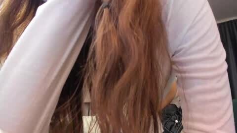 Media: Video of a person with long, wavy auburn hair, wearing a white long-sleeved top, bending forward with their hair cascading over their face. Background is blurry, suggesting an indoor setting.