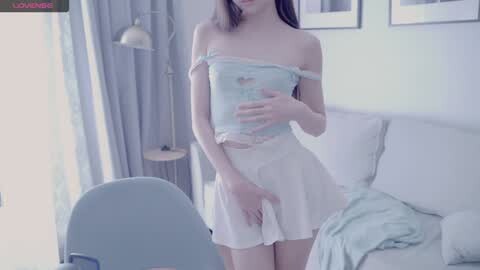 Media: Video of a slim, pale-skinned woman with long brown hair, wearing a light blue off-shoulder crop top and white mini skirt, standing in a bright, minimalist room with light curtains, a white couch, and a lamp.