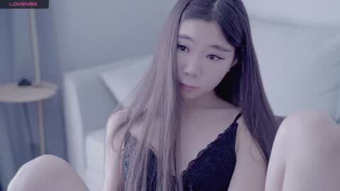 Media: A young Asian woman with long black hair and fair skin, wearing a black lace bra, sits on a bed in a softly lit room.