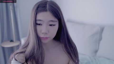 Media: A video of a young East Asian woman with long, straight brown hair, wearing a white top, sitting on a bed in a softly lit, minimalist bedroom.