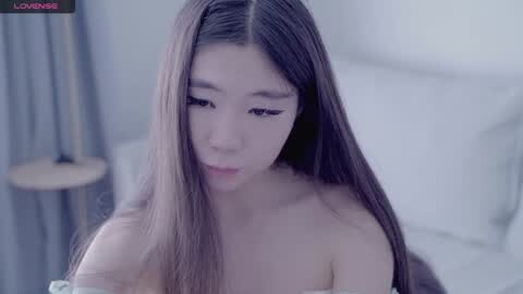 Media: Video of an East Asian woman with long, straight black hair, wearing off-shoulder attire, in a softly lit, minimalist bedroom setting.