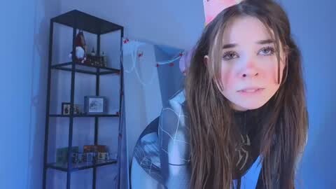 Media: Video of a young woman with long brown hair, wearing a white lab coat, standing in a room with a blue color theme.