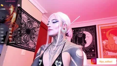 Media: Video of a pale-skinned woman with long platinum blonde hair, wearing gothic black and white lingerie, adorned with elaborate tattoos and jewelry. She stands in a room with wall art and a TV screen displaying \"Lowrider.\