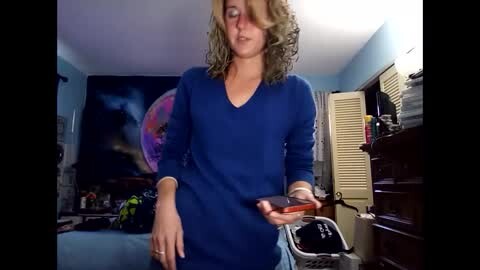 Media: A candid video of a woman with curly blonde hair in a blue V-neck sweater holding a red phone in a dimly lit, cluttered bedroom with a dark wall mural.