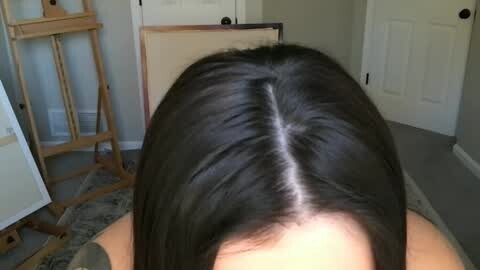 Media: A video showing a woman's dark brown, straight hair parted down the center, viewed from behind, in a home setting with light walls and wooden furniture.