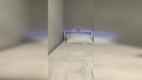 Media: Video of a minimalist, modern bedroom with a single bed against a white wall, featuring a white duvet, pillow, and a small lamp emitting soft blue light on the bedside table.