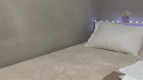 Media: Video of a minimalist bedroom featuring a single bed with beige sheets, a white pillow, and a small, clear glass lamp emitting soft blue light on the bedside table. The walls are smooth and painted in a neutral grey tone.