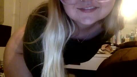 Media: A video of a smiling, light-skinned woman with long, blonde hair wearing glasses and a black shirt, leaning over. Background shows a cluttered room with a lamp, a sink, and toiletries.