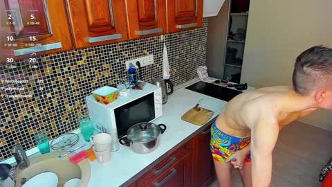 Media: Video of a shirtless man in colorful underwear, standing in a modern kitchen with wooden cabinets, tiled backsplash, and various cooking items on the counter.
