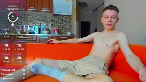Media: Video of a slim, shirtless young man with short, light brown hair, wearing beige shorts, reclining on a bright orange sofa in a modern kitchen with wooden cabinets and a tiled backsplash.