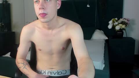 Media: Video of a shirtless, young, slender, fair-skinned man with a small penis, wearing Calvin Klein underwear, sitting in a dark room with floral decorations and a bed in the background.