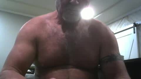 Media: Video of a shirtless, middle-aged man with a large beard, wearing a bandage on his right arm. He has a muscular build, tan skin, and a smile. The background shows a dimly lit room with a window and a ceiling light.