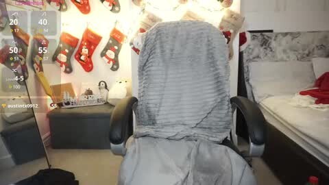 Media: Video of a woman sitting in a gray chair, wearing a gray knit scarf, in a cozy, Christmassy room with a bed, decorated wall, and hanging stockings.