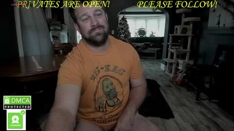 Media: A video of a bearded, muscular white man with short hair wearing an orange \"The Legend of Zelda\" t-shirt, sitting on a wooden floor, in a dimly lit, cluttered room with a cat tower, Christmas tree, and a couch.