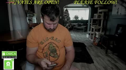 Media: Video of a bearded man in an orange t-shirt, sitting on a wooden floor, holding a phone, with a \"Private\" sign on a door in the background.