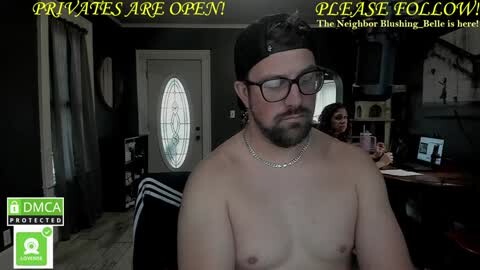 Media: Video of a shirtless, bearded man with glasses and a backward baseball cap, wearing a silver necklace, sitting in a dimly-lit room with dark walls, a white door, and a computer desk with a monitor in the background.