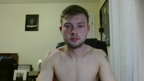 Media: Video of a shirtless, fair-skinned, young man with short brown hair and a trimmed beard, sitting indoors. He has a tattoo on his right shoulder. Background shows a framed picture and a white curtain.