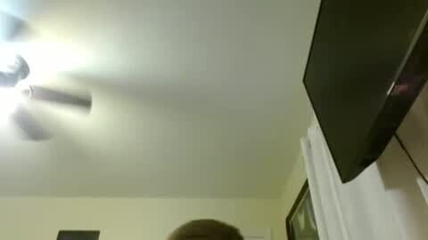 Media: Video of a dimly lit bedroom ceiling with a ceiling fan and a flat-screen TV mounted on the right wall. A person's head is partially visible, suggesting a close-up perspective.