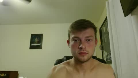 Media: Video of a young, shirtless, fair-skinned man with short brown hair, gazing directly at the camera. Background features a framed poster, a window, and a ceiling fan.