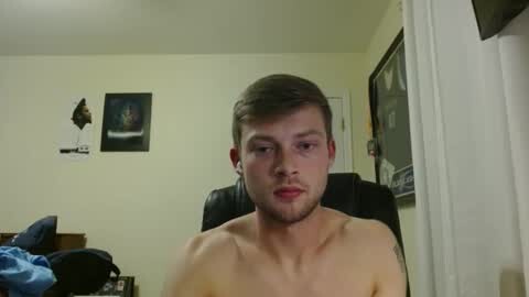Media: Video of a shirtless, fair-skinned young man with short brown hair, sitting in a dimly lit room with beige walls. Background includes a framed picture, a black chair, and a partially visible bed.