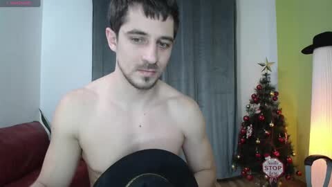Media: Video of a shirtless young man with short dark hair, fair skin, and a faint beard, wearing a black hat, in a festive room with a Christmas tree and a \"STOP\" sign.