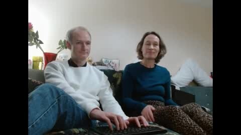 Media: Video of an elderly couple, a man with short gray hair in a white sweater and jeans, and a woman with shoulder-length brown hair in a blue sweater and leopard-print pants, sitting on a couch in a beige room, watching TV.