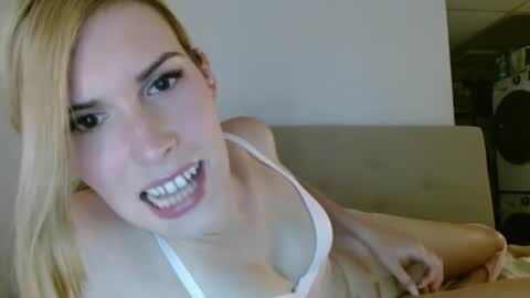 Media: A video of a young blonde woman with fair skin, wearing a white tank top, lying on a beige couch, with a surprised expression, her mouth slightly open.