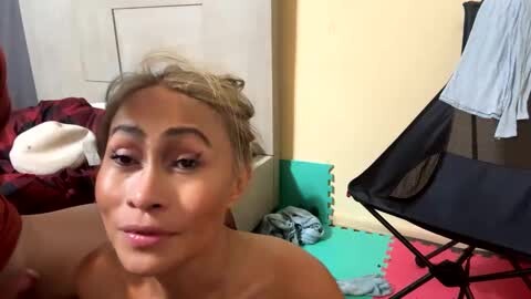 Media: A candid video of a smiling, light-skinned woman with blonde hair, wearing makeup, in a casual, cluttered room with a white door, clothes, and a black folding chair.