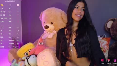 Media: Video of a woman with long black hair, wearing a black top, smiling while surrounded by plush toys, including a large teddy bear, in a dimly lit room.