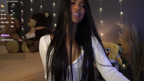 Media: A video of a smiling, light-skinned woman with long black hair, wearing a white top, in a dimly lit room with teddy bears, fairy lights, and a SpongeBob plush toy.