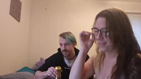 Media: A video shows a light-skinned woman with long brown hair and glasses, wearing a white top, and a man with blonde hair and glasses in a cozy, dimly lit room.