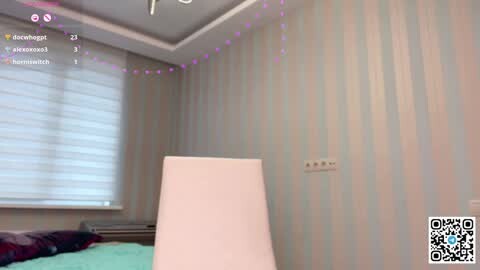 Media: Video of a modern, pastel-toned bedroom with light blue vertical-striped wallpaper, a white lampshade, and a bed with a green cover. A white window with closed blinds and a purple string of fairy lights hangs in the background.