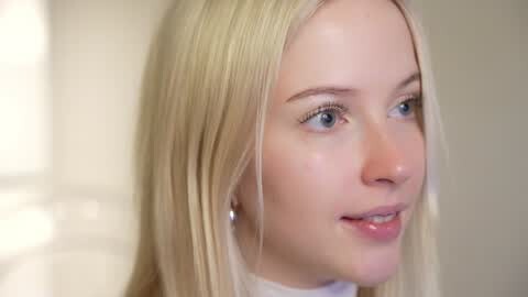 Media: Video of a fair-skinned woman with long, straight blonde hair, wearing a white top, looking to the side with a soft smile. Background is a blurred, neutral-toned room.